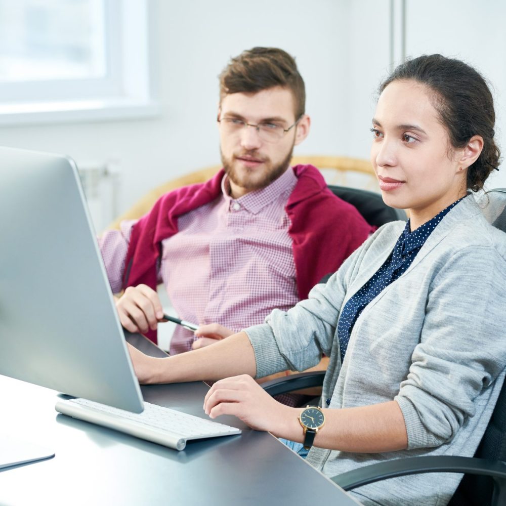 Young female office worker looking at computer screen attentively while sitting at office table with bearded male colleague